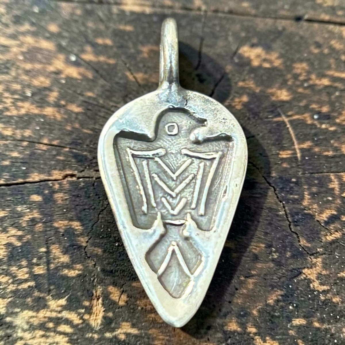 MILWORKS teardrop shaped silver charm featuring an engraved Thunderbird motif on a dark wooden surface.