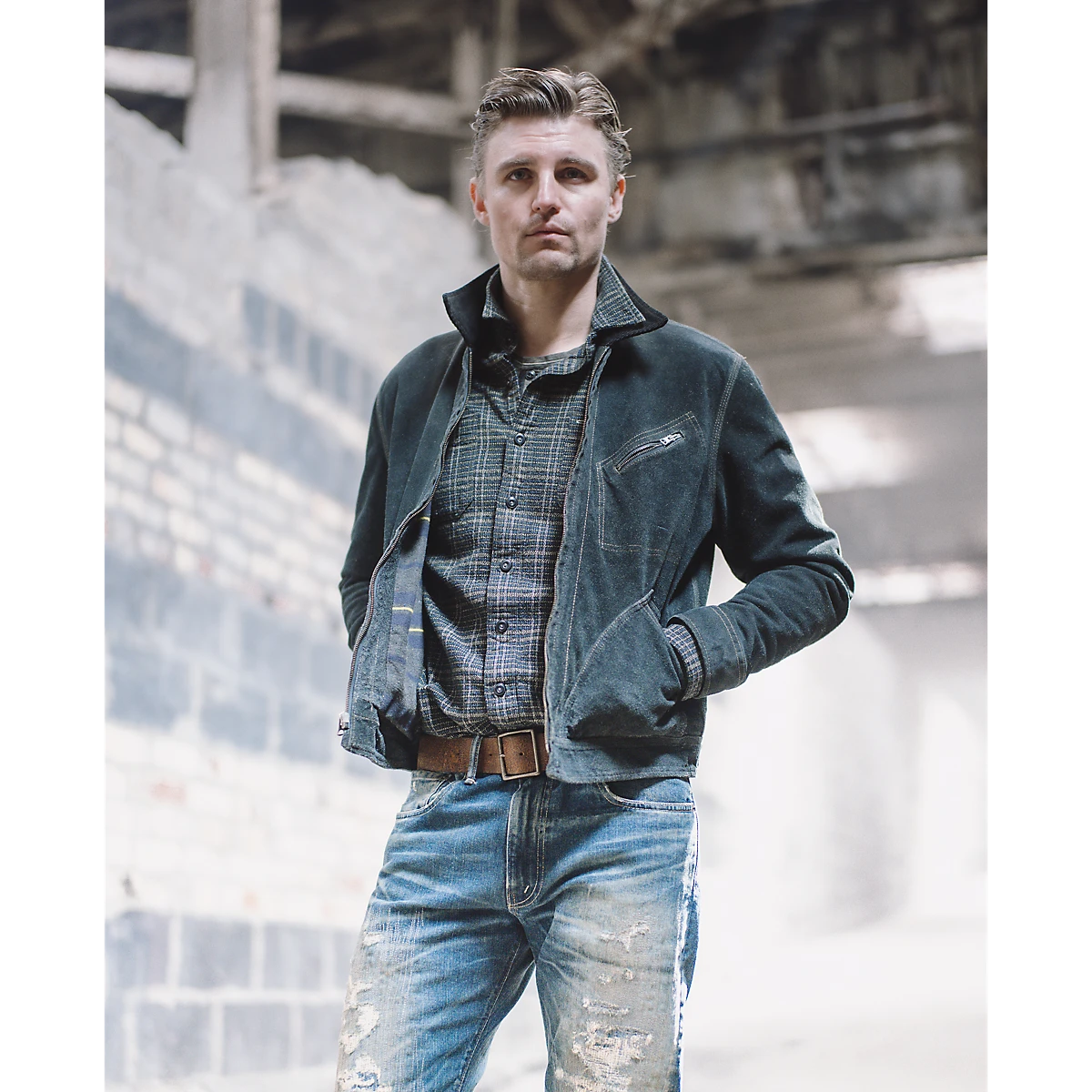 RRL model wearing a blue and green plaid cotton workshirt layered under a dark jacket and distressed jeans