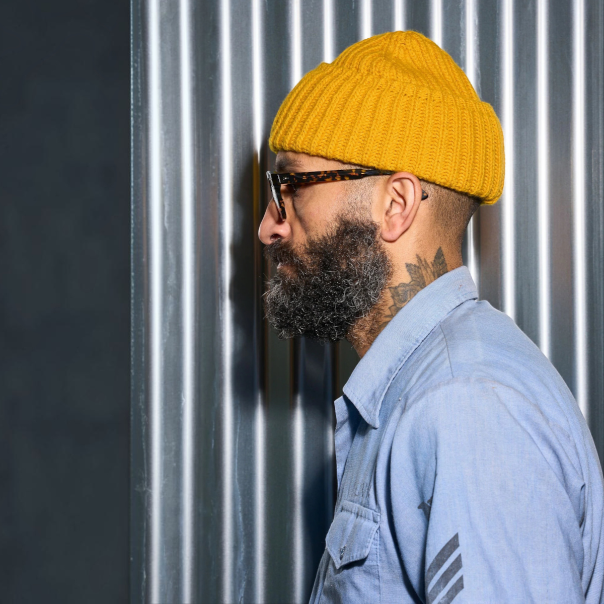milworks.momusteau watch cap in old gold lambswool worn by a bearded man in profile against a corrugated metal wall
