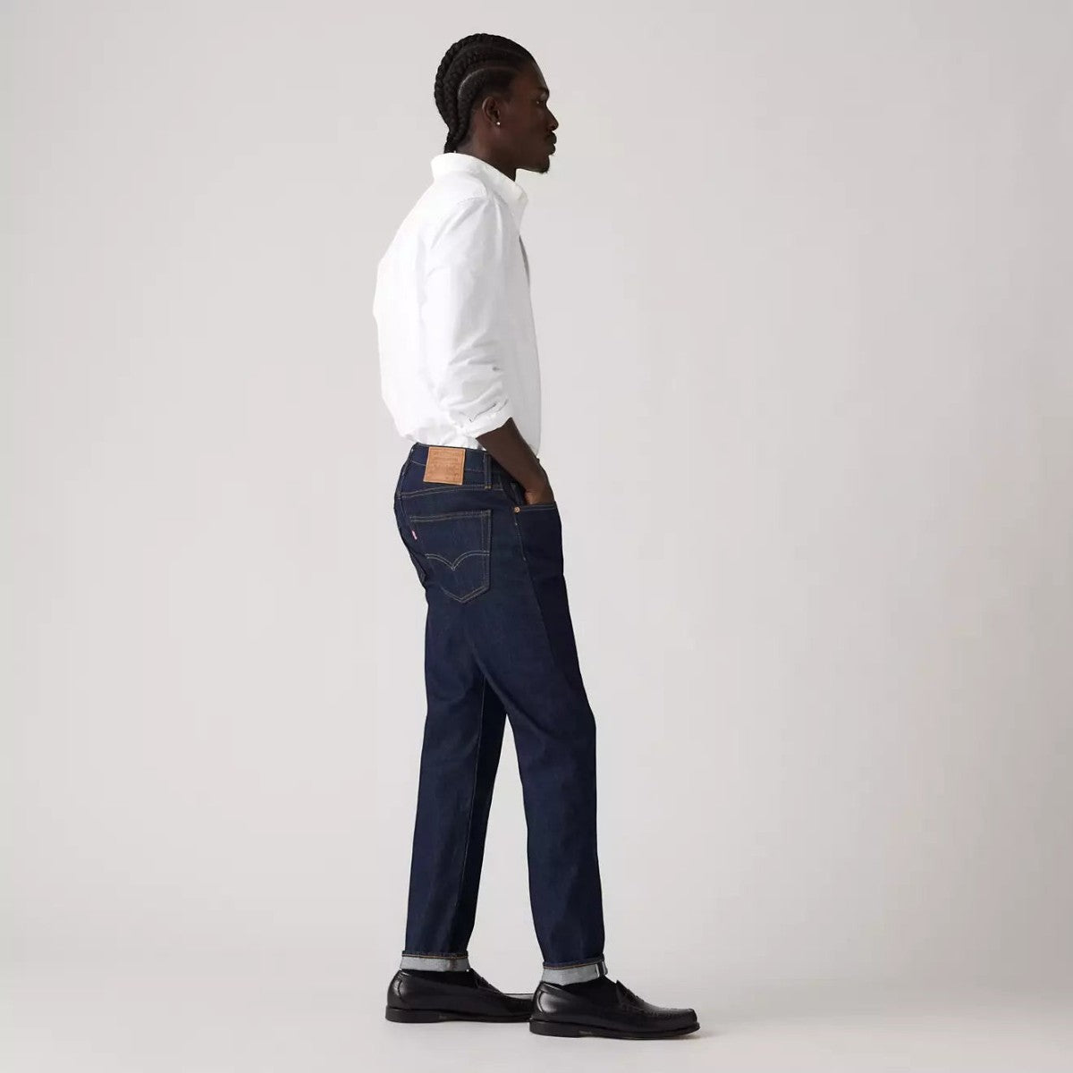 MILWORKS model wearing dark wash 502 Taper Fit selvedge denim jeans with a white shirt and black shoes, side view.