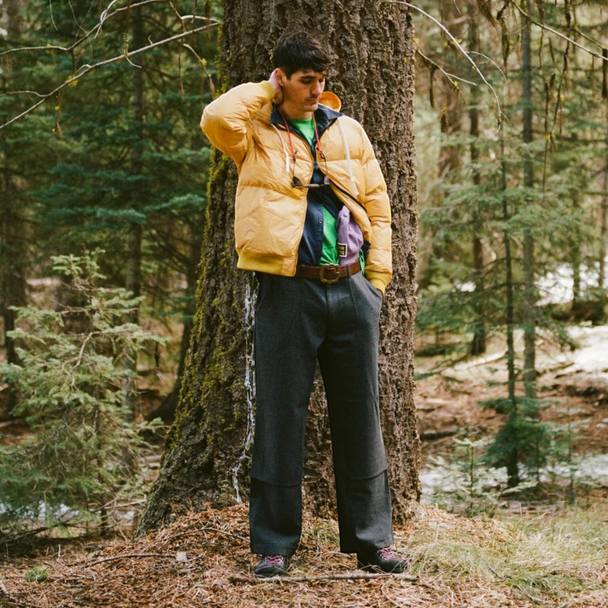 A man wearing a MILWORKS Alpine Jacket in mango nylon with a drawstring hood, standing in a forest.