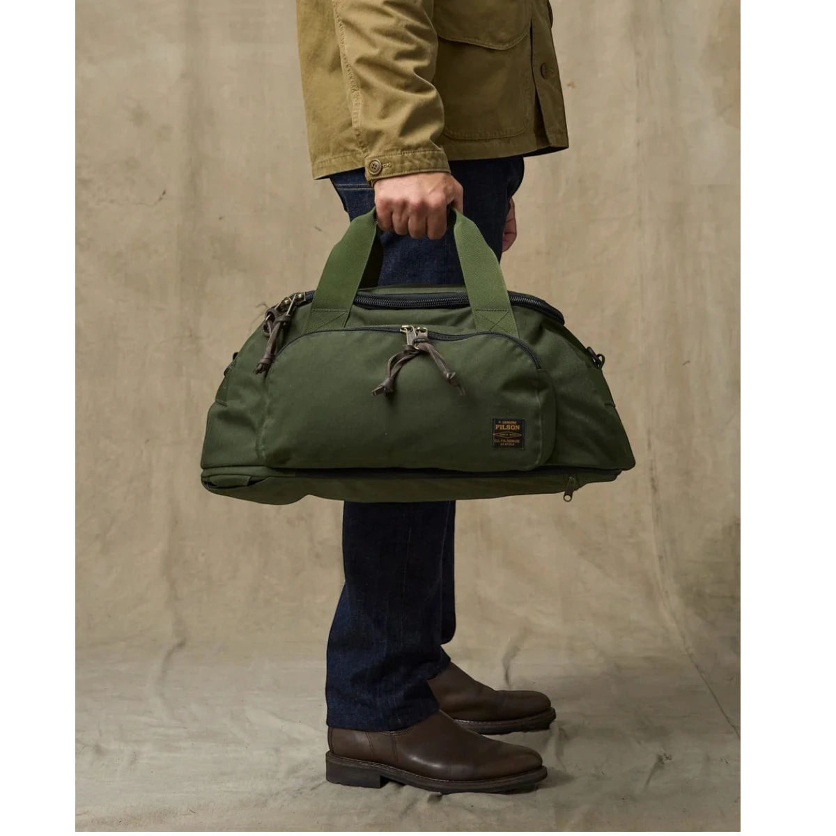 MILWORKS Dryden Duffle Pack in otter green 1000D nylon held by a person wearing a tan jacket and dark jeans