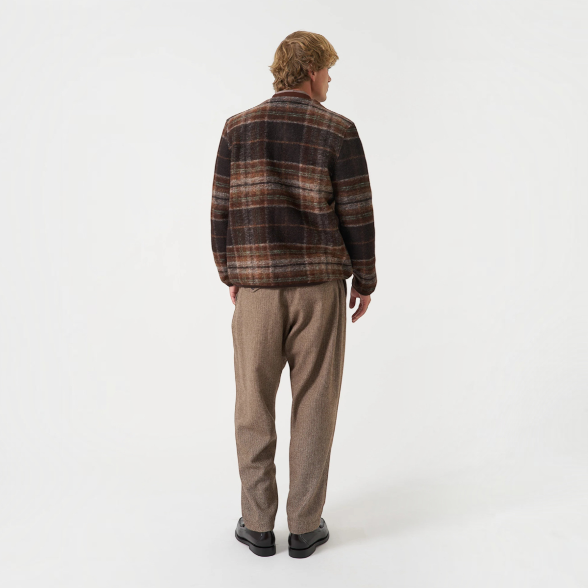 A model seen from behind wearing a MILWORKS Pino wool fleece cardigan in a brown plaid pattern with trousers.